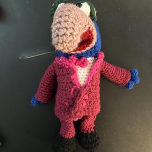 Pu&ograve; includere: Bambola all'uncinetto fatta a mano di un personaggio simile a un uccello che indossa un abito magenta con camicia bianca e papillon rosa. L'uccello ha un becco marrone, testa blu e bocca rossa. La bambola ha scarpe nere e mani blu.