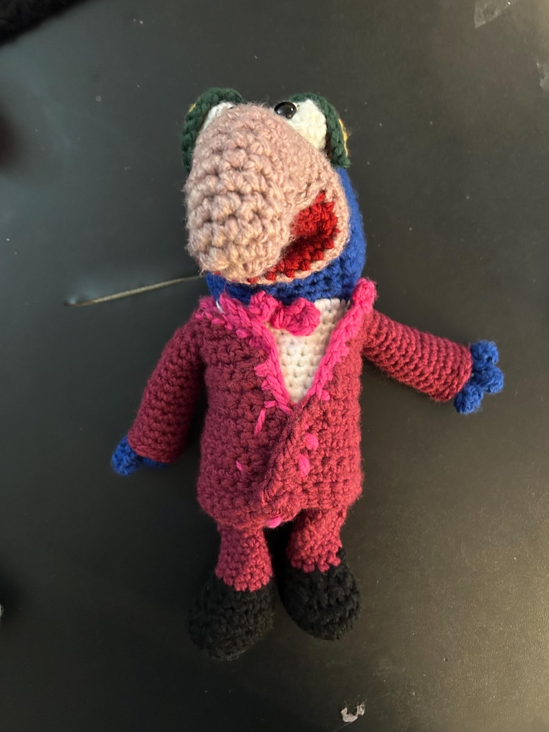 Pu&ograve; includere: Bambola all'uncinetto fatta a mano di un personaggio simile a un uccello che indossa un abito magenta con camicia bianca e papillon rosa. L'uccello ha un becco marrone, testa blu e bocca rossa. La bambola ha scarpe nere e mani blu.