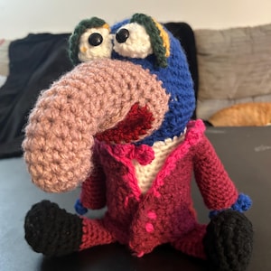 Pu&ograve; includere: Bambola all'uncinetto fatta a mano con testa blu, grande naso rosa e occhi neri. Indossa una giacca magenta con bottoni rosa e scarpe nere. Le orecchie della bambola sono verdi e gialle.