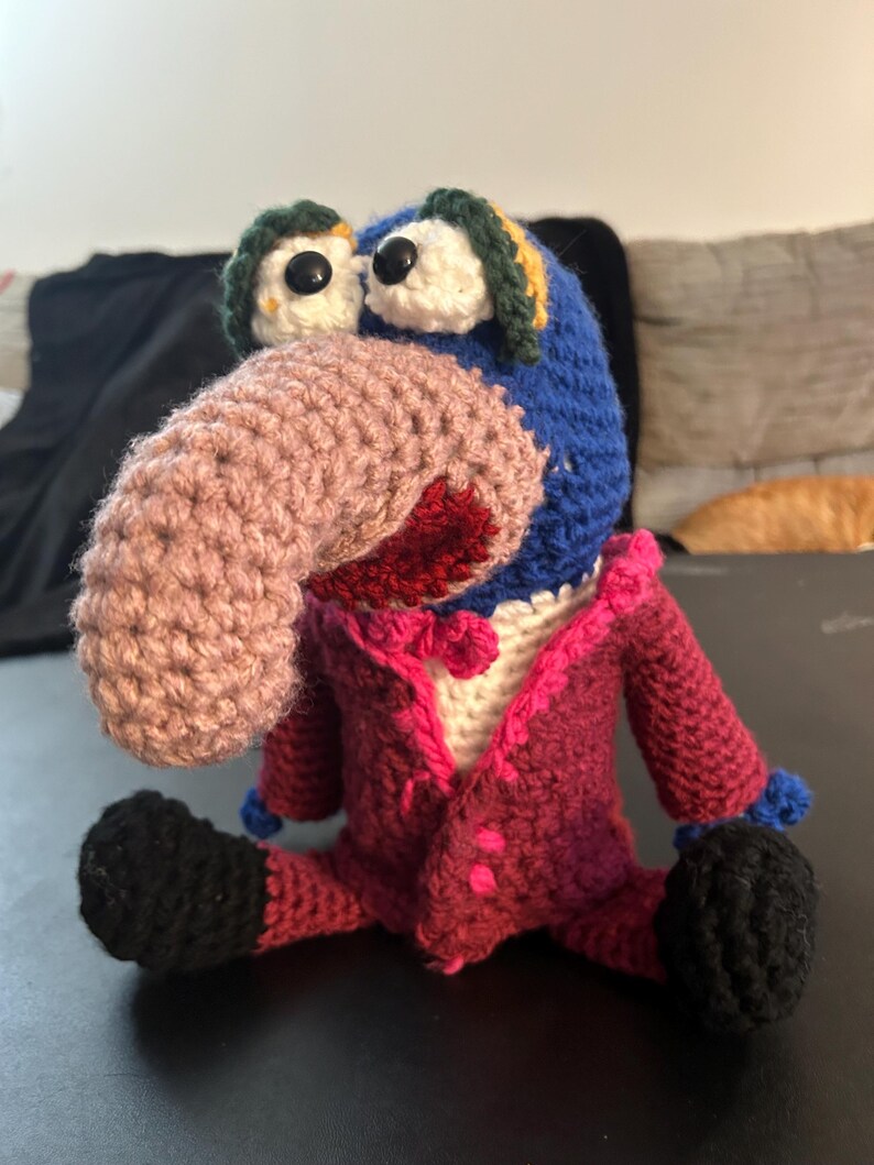 Pu&ograve; includere: Bambola all'uncinetto fatta a mano con testa blu, grande naso rosa e occhi neri. Indossa una giacca magenta con bottoni rosa e scarpe nere. Le orecchie della bambola sono verdi e gialle.