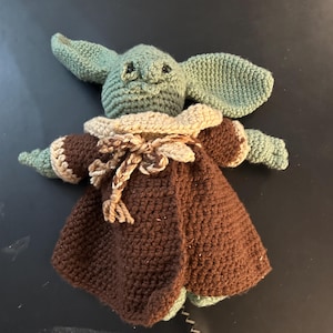 May include: Crocheted doll of a character with large ears, green skin, and a brown cloak. The doll has a tan collar and a braided rope closure. The doll is handmade and appears to be a character from a popular science fiction franchise.