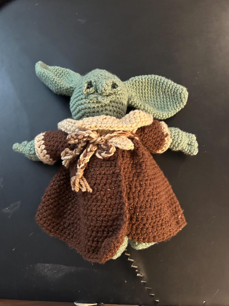 May include: Crocheted doll of a character with large ears, green skin, and a brown cloak. The doll has a tan collar and a braided rope closure. The doll is handmade and appears to be a character from a popular science fiction franchise.