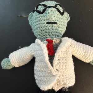 Pu&ograve; includere: Bambola all'uncinetto di uno scienziato con testa azzurra, occhiali neri e camice bianco. La bambola ha una cravatta rossa, camicia grigia e pantaloni neri. Le braccia sono tese.