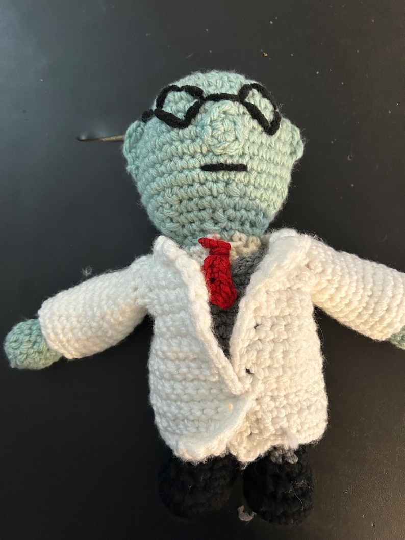 Pu&ograve; includere: Bambola all'uncinetto di uno scienziato con testa azzurra, occhiali neri e camice bianco. La bambola ha una cravatta rossa, camicia grigia e pantaloni neri. Le braccia sono tese.