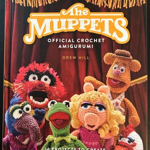 Pu&ograve; includere: Copertina di un libro all'uncinetto intitolato "The Muppets Official Crochet Amigurumi" di Drew Hill. Presenta versioni all'uncinetto dei personaggi dei Muppet, tra cui Kermit, Miss Piggy e Fozzie Bear. Il libro promette 16 progetti.