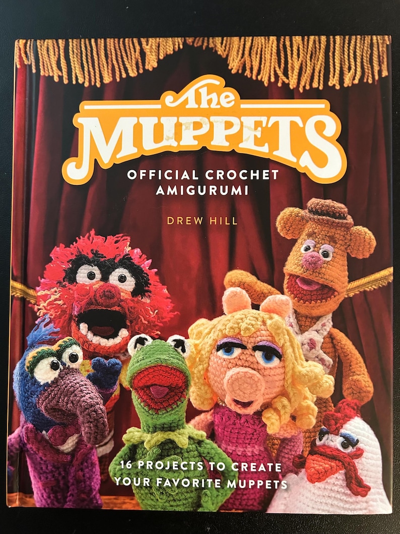Pu&ograve; includere: Copertina di un libro all'uncinetto intitolato "The Muppets Official Crochet Amigurumi" di Drew Hill. Presenta versioni all'uncinetto dei personaggi dei Muppet, tra cui Kermit, Miss Piggy e Fozzie Bear. Il libro promette 16 progetti.