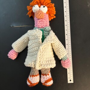 Pu&ograve; includere: Bambola all'uncinetto di un personaggio con capelli arancioni, grandi occhi bianchi e naso e bocca rosa. La bambola indossa un camice da laboratorio color crema, una camicia verde e scarpe rosa e arancioni. Un righello &egrave; accanto alla bambola.