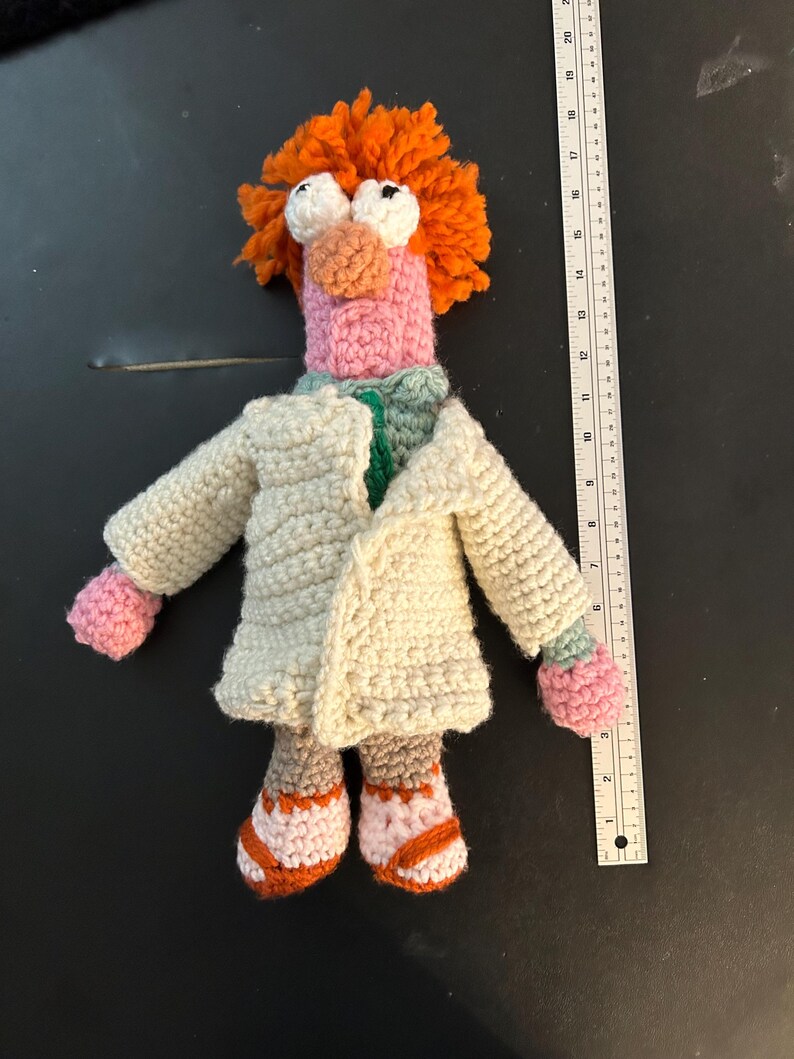 Pu&ograve; includere: Bambola all'uncinetto di un personaggio con capelli arancioni, grandi occhi bianchi e naso e bocca rosa. La bambola indossa un camice da laboratorio color crema, una camicia verde e scarpe rosa e arancioni. Un righello &egrave; accanto alla bambola.