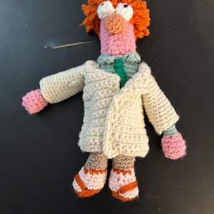 Pu&ograve; includere: Bambola all'uncinetto fatta a mano di un personaggio dei Muppet. La bambola ha capelli arancioni, grandi occhi bianchi e un naso e un viso rosa. Indossa un camice da laboratorio color crema, un colletto verde e bianco e scarpe arancioni e marroni.