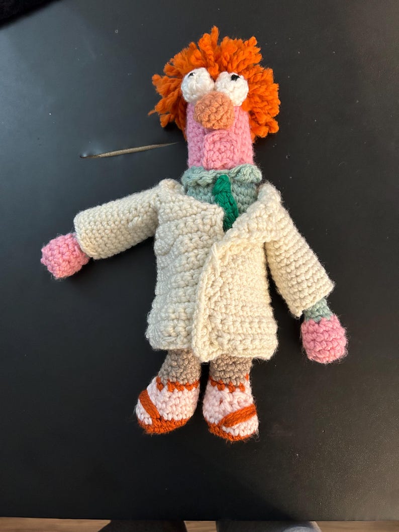 Pu&ograve; includere: Bambola all'uncinetto fatta a mano di un personaggio dei Muppet. La bambola ha capelli arancioni, grandi occhi bianchi e un naso e un viso rosa. Indossa un camice da laboratorio color crema, un colletto verde e bianco e scarpe arancioni e marroni.