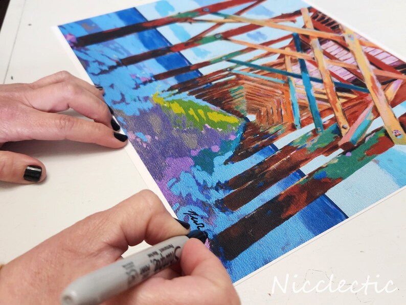 以下が含まれることがあります： 青い水を背景に、木製の桟橋を描いたカラフルな絵画。桟橋は茶色、オレンジ、ピンク、青の色合いで描かれている。絵画の右下隅には、画家のサインが見える。