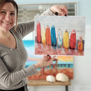 May include: A watercolour painting of a row of colourful surfboards lined up on a beach. The surfboards are painted in shades of red, orange, yellow, blue, and green. The painting has a light blue background with white clouds.