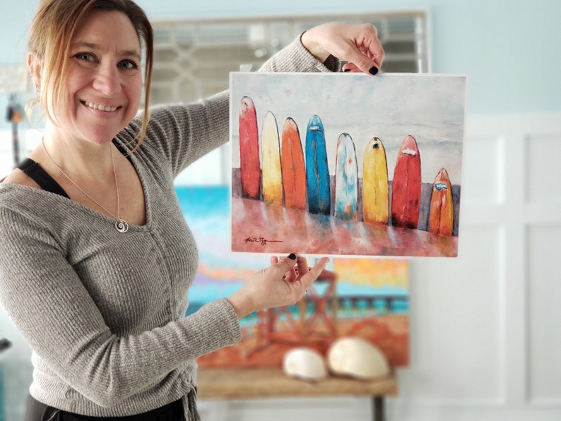 May include: A watercolour painting of a row of colourful surfboards lined up on a beach. The surfboards are painted in shades of red, orange, yellow, blue, and green. The painting has a light blue background with white clouds.
