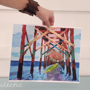 以下が含まれることがあります： 水上に伸びる木製の桟橋を描いたカラフルな絵画。桟橋は赤、オレンジ、黄色、緑、青の色合いで塗られています。水は薄い青色で、空は薄い青色で白い雲がいくつかあります。