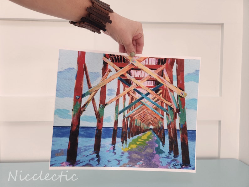 以下が含まれることがあります： 水上に伸びる木製の桟橋を描いたカラフルな絵画。桟橋は赤、オレンジ、黄色、緑、青の色合いで塗られています。水は薄い青色で、空は薄い青色で白い雲がいくつかあります。