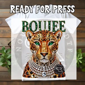 May include: A white t-shirt featuring a graphic print of a leopard with emerald green eyes, covered in sparkling rhinestones. Above the leopard, the word BOUJEE is printed in a green, gem-like font. The text READY FOR PRESS appears at the top.