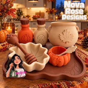 May include: A set of autumn-themed kitchenware including a brown tray, a beige leaf-shaped bowl with a brown honey dipper, a small orange pumpkin-shaped bowl with a spoon, two small bottles with pumpkin and mushroom motifs, and a beige cauldron.