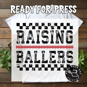 May include: White t-shirt with the text "READY FOR PRESS" at the top. The shirt features the words "RAISING BALLERS" in a distressed black font, with a red baseball stitch design. A black and white checkered pattern borders the text.
