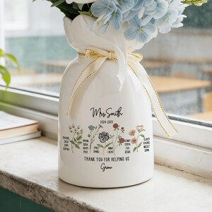 May include: A white ceramic vase filled with a bouquet of artificial flowers, including white peonies, blue hydrangeas, and daisies. The vase is tied with a gold ribbon and features the text "Mrs. Smith 2024-2025" and the phrase "Thank you for helping us Grow."