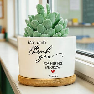 May include: A white ceramic plant pot with a succulent, on a bamboo coaster. The pot has the text "Mrs. Smith, thank you for helping me grow" with a red heart and the name "Amelia". The plant has green leaves.
