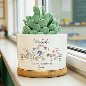 Puede incluir: Una maceta de cerámica blanca con una planta suculenta, sobre un posavasos de bambú. La maceta está decorada con ilustraciones florales y el texto "Mrs. Smith 2024-2025" y "THANK YOU FOR HELPING US Grow", junto con los nombres de los alumnos.