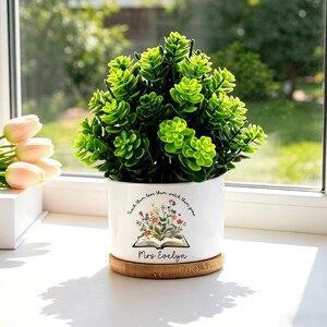 Puede incluir: Una maceta de cerámica blanca con una planta verde, sobre un posavasos de madera. La maceta tiene un diseño floral y el texto "Teach them, love them, watch them grow" y "Mrs. Evelyn".