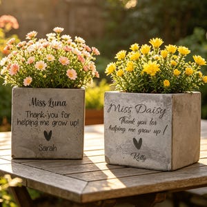 May include: Two square concrete planters with flowers. One has pink and white flowers, the other has yellow flowers. Each planter has text that reads "Thank you for helping me grow up!" and a name.