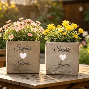 May include: Two square concrete planters with flowers, one with pink and white daisies, the other with yellow flowers. Each planter has the names "Sophia" and "James", a heart, and the date "07.26.2025".