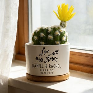 以下が含まれることがあります： 「Love grows」の文字と「Daniel & Rachel」の名前、日付「02.16.2026」が書かれた白いセラミックポットに入った小さなサボテン。 サボテンは鮮やかな黄色の花を咲かせ、木製のコースターの上に置かれています。