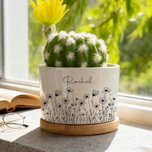 Puede incluir: Un cactus verde con una flor amarilla en una maceta de cerámica blanca. La maceta tiene un diseño floral y el nombre "Rachel" en escritura cursiva. La maceta está sobre un posavasos redondo de madera.