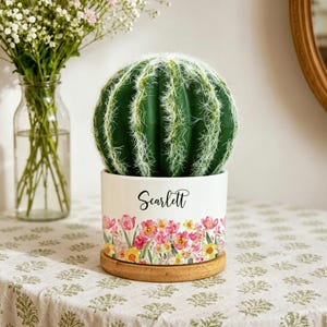 May include: A round, green, spiky artificial cactus sits in a white ceramic planter. The planter features a colorful floral pattern of pink and yellow flowers along the bottom and the name Scarlett printed in black script lettering on the front.