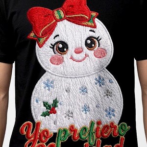 Puede incluir: Una camiseta negra con un muñeco de nieve blanco bordado que lleva un lazo rojo, mejillas rosadas y detalles de copos de nieve azules. Debajo del muñeco, el texto "Yo prefiero Navidad" está bordado en letras cursivas rojas y verdes.