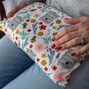 May include: A white cushion featuring a bee and floral design. The pattern includes grey beehives, yellow and black bees, pink and red flowers, and green leaves. The cushion is held on a person's lap.