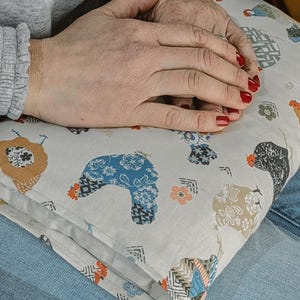 May include: A folded pillow with a repeating pattern of colourful chickens in blue, brown, and black. The pillow is resting on a person's lap. The pillow is light-coloured.