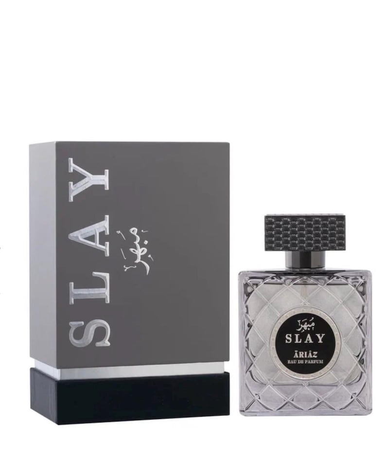 Może przedstawiać: Zestaw butelki i pudełka perfum. Pudełko jest ciemnoszare z napisem "SLAY" dużymi, srebrnymi literami. Butelka perfum jest kwadratowa z teksturowaną nakrętką.