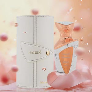May include: A white perfume bottle with a marble design and a copper-colored top sits next to a white cylindrical case. The bottle has a gold label that reads "Hamidi". The background is a soft pink with floating orbs.