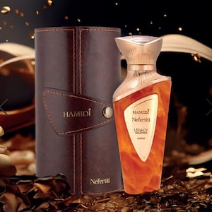 May include: A perfume bottle with a gold cap and a brown and orange body, next to a brown leather case. The bottle is labeled "HAMIDI Nefertiti LEGACY COLLECTION PARFUM". The case has the word "HAMIDI" printed on it.