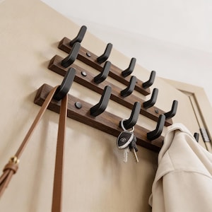 Könnte beinhalten: Ein hölzerner Wand-Kleiderhaken mit drei Reihen schwarzer Haken. Eine braune Ledertasche und eine beige Jacke hängen an den Haken. Schlüssel hängen ebenfalls an einem der Haken. Der Kleiderhaken ist an einer hellbeigen Tür montiert.