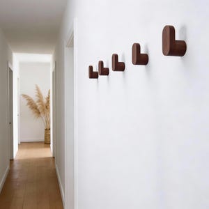 May include: A series of five wooden wall hooks in a row, each with a rounded L-shape. The hooks are a dark brown color and mounted on a white wall. The hallway has a wooden floor and a basket of dried plants.