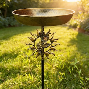 May include: A bronze bird bath featuring a butterfly and sun design. The bowl-shaped basin sits atop a black metal pole. The butterfly has orange and black markings. The bird bath is set in a garden with green grass.