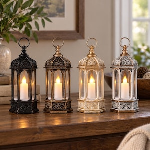 May include: Four hexagonal lanterns with intricate embossed patterns and top rings, each containing a single white LED pillar candle. The lanterns are finished in black, bronze, gold, and distressed white, standing in a row on a wooden surface.