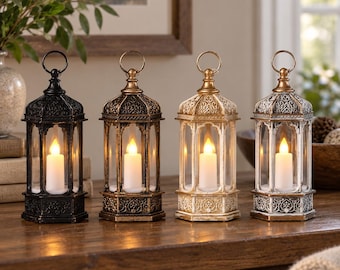 May include: Four hexagonal lanterns with intricate embossed patterns and top rings, each containing a single white LED pillar candle. The lanterns are finished in black, bronze, gold, and distressed white, standing in a row on a wooden surface.