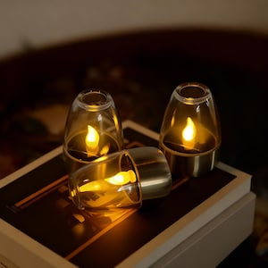 Könnte beinhalten: Drei batteriebetriebene Kerzenlampen mit warmem, flackerndem Licht. Jede Lampe hat einen transparenten Glasschirm und einen goldfarbenen Sockel. Die Lampen sind auf einer dunklen Oberfläche angeordnet und schaffen eine gemütliche Atmosphäre.