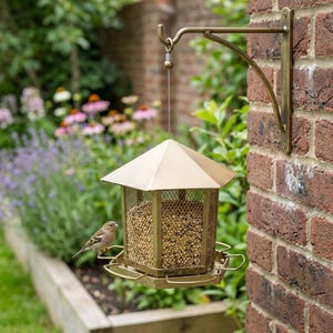 May include: A hexagonal metal bird feeder with a brass-toned finish, featuring a mesh body filled with birdseed, a pointed roof, and a circular perch. The feeder hangs from a matching curved metal wall bracket mounted on a brick surface.