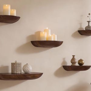 May include: Wooden floating shelves in a warm brown finish, each holding decorative items. Some shelves display white candles of varying heights, while others feature vases and decorative spheres. The shelves are mounted on a neutral-colored wall.