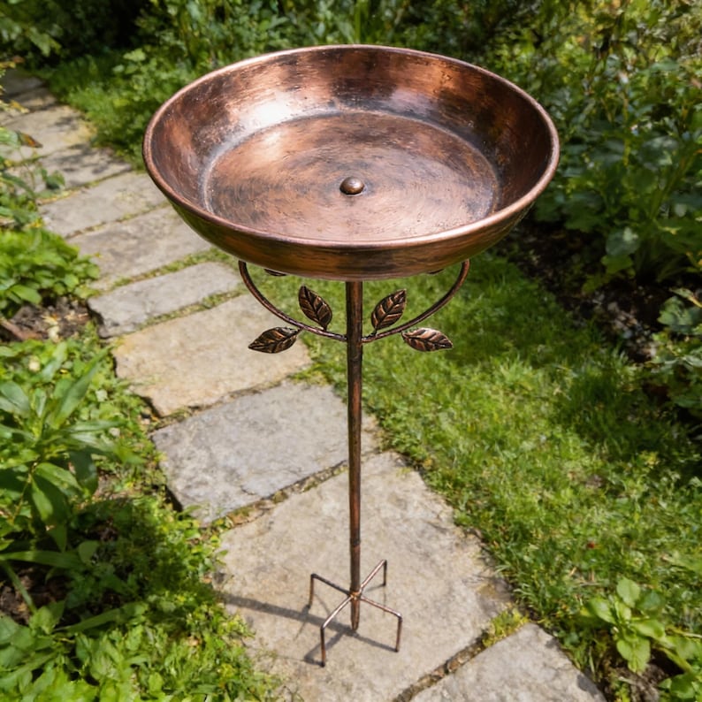 May include: A copper-colored bird bath with a round basin and a decorative leaf design. The bird bath is supported by a slender, bronze-colored stand with a four-legged base, perfect for a garden or outdoor space.