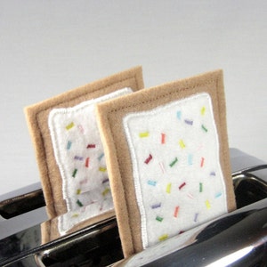 May include: Two felt fabric pop-tart style snacks with white frosting and colorful sprinkles inside a silver toaster.
