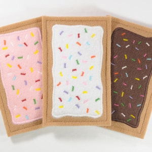 May include: Three felt squares resembling a popular breakfast pastry. The squares are brown, pink, and white with colorful sprinkles on top.