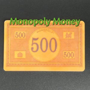 May include: A Monopoly money bill with a value of 500. The bill is orange with the text "Monopoly Money" in green at the top. The number 500 is prominently displayed in a large circle in the center.