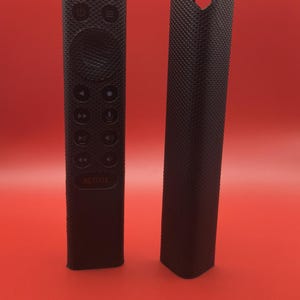 May include: Two black remote control covers. One cover is on a remote with buttons and the word "NETFLIX". The other cover is a solid rectangle with a heart-shaped cutout at the top. Both covers have a textured surface.